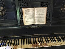 Piano droit noir, clavier jauni, revêtement ivoire défectueux, partition sur pupitre