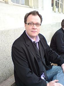 一個戴眼鏡的男子，身穿黑夾克、背心和領結、粉襯衫和牛仔褲，坐在大理石花紋的牆壁前。