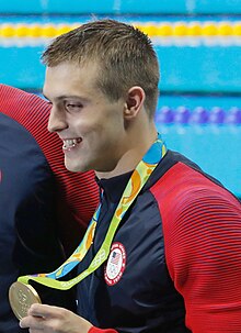 Ryan Held in blaurotem Trainingsanzug bei der Siegerehrung 2016, mit der rechten Hand zeigt er den Fotografen seine Goldmedaille.