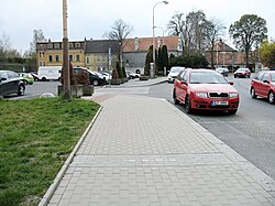 Pohled na zcela změněné centrum někdejší židovské čtvrti v České Lípě. V pozadí před renesančním Červeným domem je parkoviště a parčík s pomníkem v místech, kde stávala synagoga. Místo domu čp. 692, který byl, stejně jako řada dalších, zbořen na počátku 80. let 20. století, je jen trávník (vlevo). V roce 2018 zde byly do chodníku vsazeny tzv. Stolpersteine neboli kameny zmizelých (na snímku v popředí), připomínající čtyři členy rodiny Wolffovy z čp. 692, kteří zahynuli v koncentračním táboře.