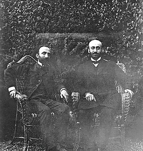 Photographie en noir et blanc de deux hommes barbus assis sur des chaises.