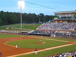 A green baseball diamond illuminated by lights at dusk with players on the field and people watching from the grandstand