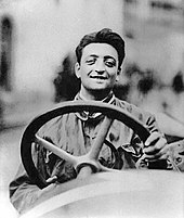 Photo d'Enzo Ferrari, souriant, derrière le volant d'une voiture.