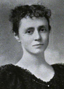 A young white woman with dark hair and eyes, wearing a dark dress with a scooped neckline