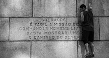 Em preto-e-branco, um rapaz jovem se apoia no monumento à frente da frase "Soldados! É fácil a missão de commandar homens livres: basta mostrar-lhes o caminho do dever!".