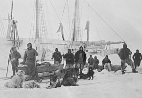 Группа мужчин позируют на льду с собаками и санями, на заднем плане виден контур корабля.