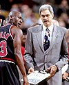 Man in gray hair, glasses, and suit on the edge of a basketball court and beside a player in uniform