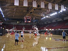 Vue de l'intérieur d'une salle de basket-ball dans laquelle les sièges des tribunes sont rouges. Des maillots blancs sont accrochés au plafond.