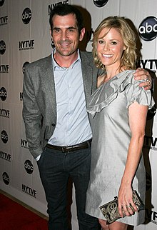 A blonde woman wearing a silver dress smiles as she stands on a red carpet to the left of a man with dark hair wearing a blue striped shirt, gray blazer, and black pants. They are both standing in front of a backdrop with „NYTVF“ and a logo that is a round black ball with the letters „abc“ on it