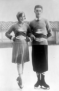 A woman and a man in figure skating blades stand on an outdoor ice rink posing for a shot. On the left, the woman has both hands in her waist, while the man has his right arm around her left arm.