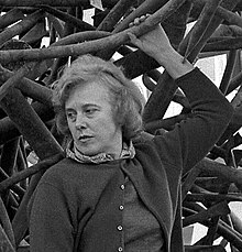 Claire Falkenstein, standing on one of her sculptures, her left arm raised above her head holding part of the sculpture