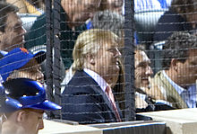 Трамп на бейсбольном матче в июле 2009 года. Он одет в бейсболку и сидит среди большой толпы за защитной сеткой.