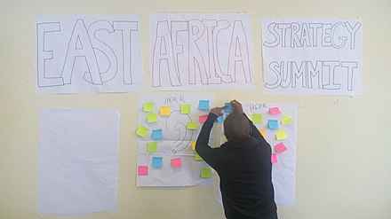 Participant is attaching sticky notes onto flip chart paper attached to a wall. Above that is a hand draw sign on flip chart paper that reads 'East Africa Strategy Summit'