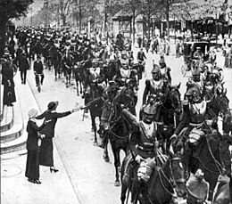 Černobílá fotografie ukazuje velký oddíl vojáků jedoucích na koních ulicí. V popředí žena stojící poblíž obrubníku, která jednomu z projíždějících vojáků cosi podává.