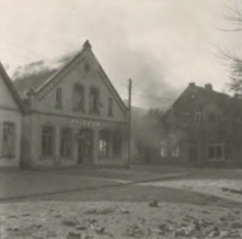 Photographie en noir et blanc montrant un bâtiment avec de la fumée et des flammes s'échappant du toit et de l'intérieur. Sur la façade est écrit "Friseur", signifiant "coiffeur" en allemand.