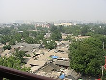 Вид на остроконечные крыши одноэтажных домов с вкраплениями деревьев на переднем плане. На заднем плане, слегка скрытые смогом, видны более высокие и современные здания.