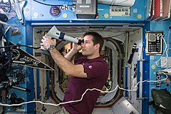 Thomas Pesquet w stroju codziennym w module Destiny wykonuje rutynowe badanie dna oka przy użyciu oftalmoskopu