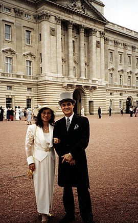 A colour photograph of Jason Tsai, alongside his wife Ruth Tsai. The couple are dressed in formal wear: Jason is wearing a black morning suit with a grey top hat and matching grey necktie and pocket handkerchief; his wife Ruth is wearing a full-length white dress, matching white jacket and white hat. The couple are standing in the red-gravelled courtyard of Buckingham Palace - the central London official residence of Queen Elizabeth II, the reigning monarch of the United Kingdom of Great Britain and Northern Ireland. In the background are other unidentifiable people in similar attire.