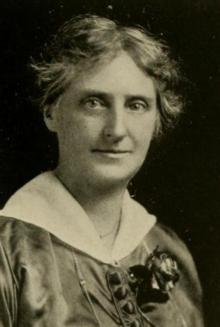 A middle-aged white woman with grey hair, wearing an academic gown with a corsage pinned to the front