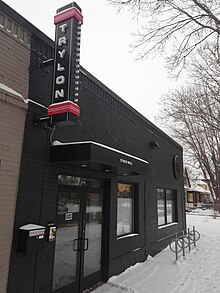 A black storefront with a vertical sign reading "TRYLON" projecting from high on the wall. There is snow on the ground and a set of three bike racks in front of the cinema.