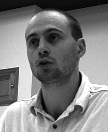 black-and-white image of Yves Engler wearing a light shirt, looking right of camera and appearing to speak
