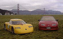 Two sports cars, the yellow one on the left, and a red one on the right, parked on a large, grassy plain. In the background stands the Golden Gate Bridge.