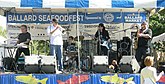 Four people on a stage; from left to right, they are playing a keyboard, a trumpet, a drum set, a guitar, and a saxophone. In the background are various signs, and on the stage are microphone stands and other audio equipment.