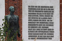 Eine Bronzestatue zeigt einen abgemagerten Mann und steht vor einer weiß gestrichenen Fassade, an die mit Großbuchstaben eine fünfzehnzeilige Inschrift angebracht ist