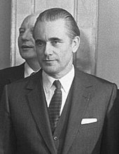 Buste d'un homme portant le costume cravate, en noir et blanc, une deuxième personne est masquée derrière lui.