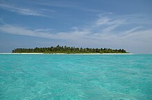 Daytime edge-on view of a small forested island amid a shallow turquoise lagoon. The dark sky blue above is streaked by thin, wispy clouds of various sizes. The lone island is evenly low-slung, no part of it reaching any substantial height whatever.