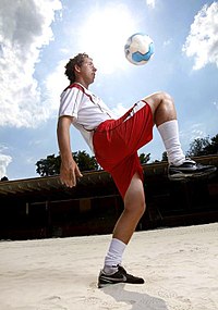 Мужчина стоит на одной ноге, при этом колено другой ноги поднято до пояса. Мяч можно увидеть в воздухе примерно на 50 см выше его колена.