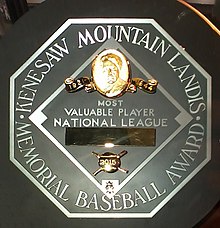 A black circle with an octagonal silver plaque in the middle. The edge of the plaque reads "KENESAW MOUNTAIN LANDIS MEMORIAL BASEBALL AWARD". In the middle of the octagon is a baseball diamond which contains, from the top, Judge Landis' face in gold, "Most Valuable Player", the winner's league, his name in a gold rectangle, and his team.