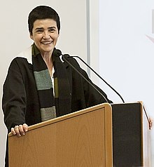 Cliché de Simone Bonnafous, une femme blanche aux cheveux noirs et courts, vêtue de noir, debout derrière un pupitre, souriant et regardant en biais vers l'appareil photo.