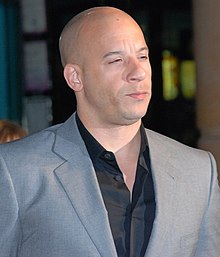A bald, multi-racial man in a gray suit. Turned at a 45 degree angle from the camera, he squints in the direction of an off-camera light source, which illuminates his face.