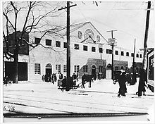 Exterior of the arena's entrance with people arriving by horse-drawn carriages and on foot
