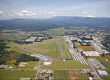 Ein Flugplatz mit drei winkligen Landebahnen, umgeben von Hangars, Lagerhäusern und offenem Grasland. Berge und Wälder sind im Hintergrund zu sehen.