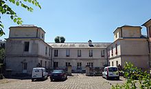 Au premier plan, la cour pavée ; au centre l’ancienne maison du domaine de la Chambre. Bâtiment blanc en forme de U, décoré de deux corniches, et au toit en ardoise.