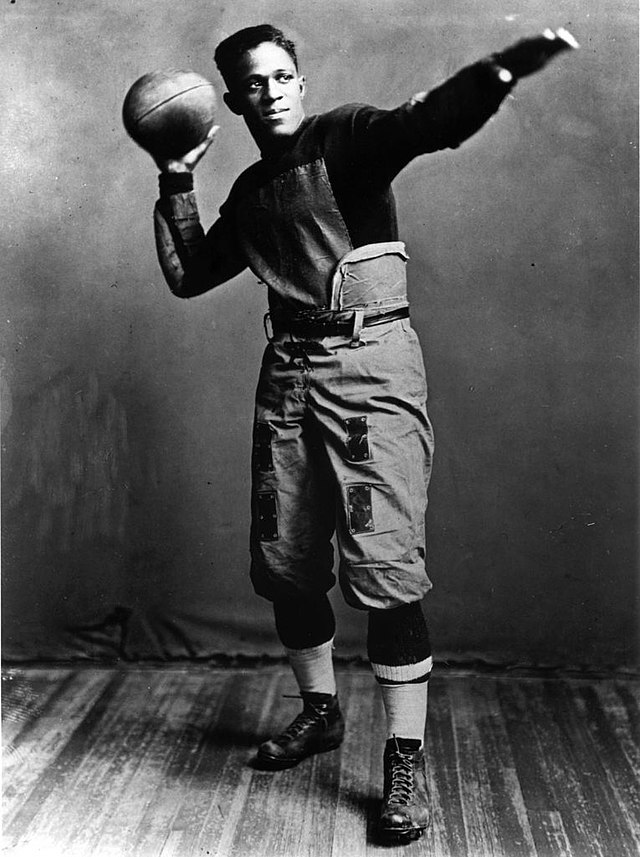 Pollard in football uniform staring off in the distance and striking a classic football pose; one arm is outstretched, while the other holds a football, as if he were about to throw it.