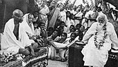 Photo of a formal function, an aged bald man and old woman in simple white robes are seated side-by-side with legs folded on a rug-strewn dais; the man looks at a bearded and garlanded old man seated on another dais at left. In the foreground, various ceremonial objects are arrayed; in the background, dozens of other people observe.