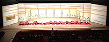 Quatorze pessoas vestidas em roupas típicas japonesas em reverência ao público no palco de um teatro.
