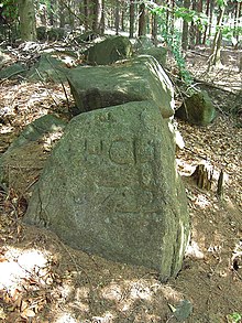 Uprostřed velký tmavě šedý kámen nepravidelného tvaru s vyrytým nápisem +HCL 1722, od něj směrem do pozadí se táhne tměvý kamenný val. Kolem kamene i valu je les, v popředí hrabanka, v pozadí jsou vidět části stromů.
