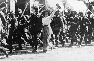 Černobílá fotografie ukazuje skupinu kráčejících vojáků. Vojáci vypadají vesele a nesou pušky s bajonety. Uprostřed obrázku se s jedním vojákem vede žena oblečená ve světlých šatech s širokým kloboukem.
