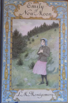 A rectangular image used for the book cover of the novel "Emily of New Moon". At the centre of the image is Emily Starr wearing a pink dress, covered by a grey coat. She is clasping her hands over her heart. She is wearing a blue bonnet on her head and standing outdoors on the base of a small hill covered in pine trees. Surrounding this painting is a blue border with intricate floral decor and brass decoration that twist to frame the title at the top.