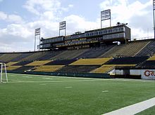 Photo of a green turf field in the foreground, and a single-tiered stadium in the background with black and gold-painted seats