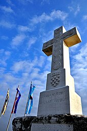 Вид на большой каменный крест с хорватским гербом. На фоне голубого неба выделяются крест и три вертикально подвешенных флага.