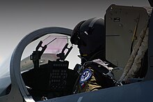 Three-quarter hind view of jet fighter cockpit. A helmet-cladded pilot is present, as is a head-up display in front of him