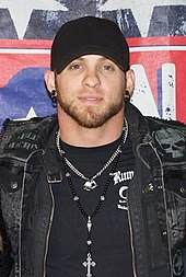 A young man with dark beard stubble, wearing a black cap, dark denim jacket, black t-shirt an various necklaces