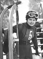 Woman holding upright a large sled with her right arm. She wears a belted dark jumpsuit with a vest over it displaying the number five, dark gloves, and a helmet with attached white-framed goggles.