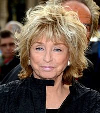 Femme de 72 ans, cheveux au balayage blond, les yeux très bleus, souriante.