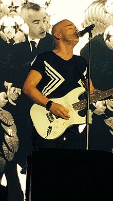 A man with short gray hair is wearing a black shirt and is playing on a white guitar